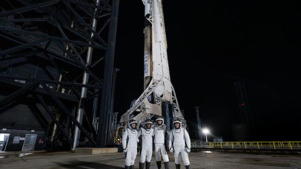 Four astronauts in white suits standing in front of a rocket at night.